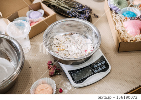Metal bowl containing white powder and botanical inclusions sits on digital scale surrounded by packaging boxes. Cosmetic raw materials, ingredient study, clean formulation, skincare transparency. Metal bowl containing white powder and botanical inclusions sits on digital scale surrounded by packaging boxes. Cosmetic raw materials, ingredient study, clean formulation, skincare transparency. 136190072