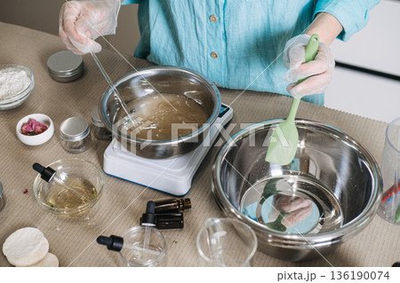 Person uses glass rod to stir liquid in metal bowl on digital scale while holding green spatula. Precise cosmetic formulation process, aesthetic beauty lab scale, artisan skincare manufacturing. 136190074