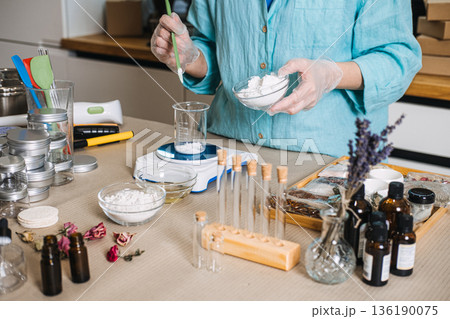 Person adds white powder from bowl into glass beaker on electronic scale surrounded by botanical ingredients. Lab-style skincare, botanical chemistry, diy cosmetic formulation, apothecary jars. Person adds white powder from bowl into glass beaker on electronic scale surrounded by botanical ingredients. Lab-style skincare, botanical chemistry, diy cosmetic formulation, apothecary jars. 136190075