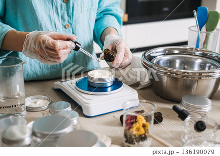 Person uses glass dropper to add oil to white cream in metal jar on digital scale. Lab-style beauty aesthetic, precise cosmetic formulation, diy apothecary lab, scientific skincare diy. 136190079