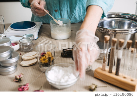 Hands in gloves reach for ingredients while mixing white powder into beaker among jars of dried flowers. botanical extracts, raw beauty ingredients, herbal apothecary, natural formulation art. 136190086