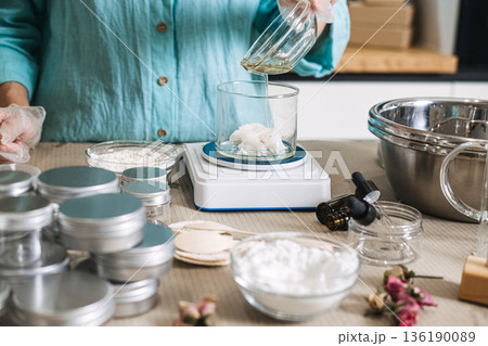 Hand in glove pours liquid oil from bowl into beaker containing white solid butter on digital scale. cosmetic formulation process, oil phase emulsion, cream texture transition, handmade moisturizer. Hand in glove pours liquid oil from bowl into beaker containing white solid butter on digital scale. cosmetic formulation process, oil phase emulsion, cream texture transition, handmade moisturizer. 136190089