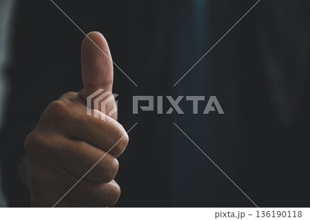 Sign of approval shows a man hand pose with a thumbs up gesture in a closeup, this positive sign communicates success and agreement against a dark professional background 136190118