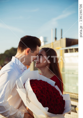 Couple shares a moment with roses in hand near a waterfront at sunset in an outdoor setting Couple shares a moment with roses in hand near a waterfront at sunset in an outdoor setting 136190786