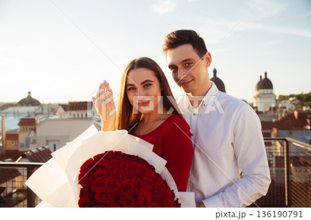 Couple shares a moment on a rooftop with a bouquet of roses during sunset in a city setting 136190791