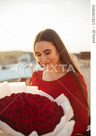 Woman smiles while holding a large bouquet of roses on a rooftop during sunset Woman smiles while holding a large bouquet of roses on a rooftop during sunset 136190802