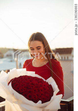 Woman holds large bouquet of red roses on balcony during sunset in city 136190803