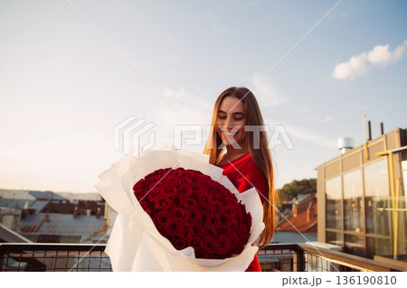 Woman holds a large bouquet of red roses on a rooftop during sunset in a city setting Woman holds a large bouquet of red roses on a rooftop during sunset in a city setting 136190810