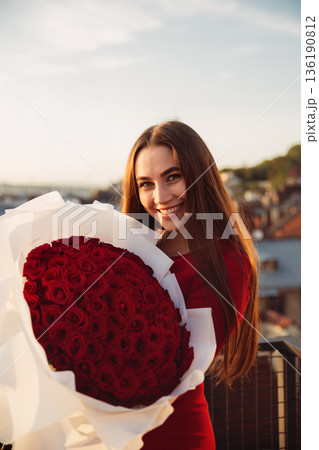 Woman smiles while holding large bouquet of red roses on balcony during sunset in city 136190812