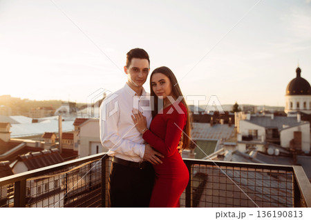 Couple stands together on a rooftop during sunset with city buildings in the background Couple stands together on a rooftop during sunset with city buildings in the background 136190813