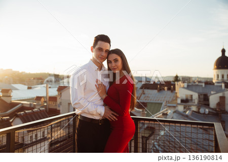 Couple stands together on rooftop during sunset overlooking city skyline Couple stands together on rooftop during sunset overlooking city skyline 136190814