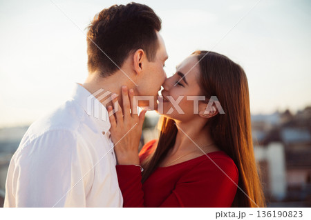 Couple shares a kiss on a rooftop during sunset in a city setting with buildings in the background 136190823
