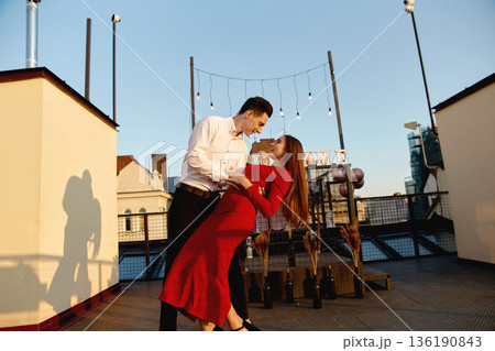 Couple dances on rooftop at sunset enjoying city view while celebrating their love with string lights around them 136190843