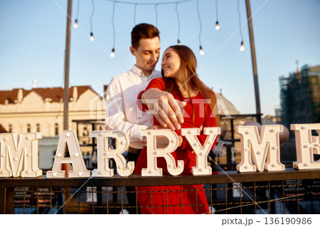 Couple celebrates engagement with a creative proposal on a rooftop at sunset surrounded by decorative lights 136190986