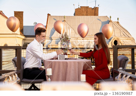 Couple enjoys a special dinner on a rooftop terrace during sunset with balloons and candles in the background 136190987