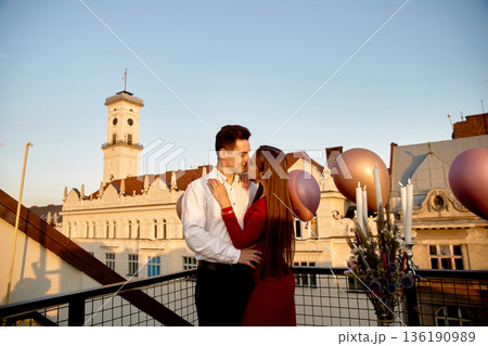 Couple shares a moment on a rooftop with balloons during sunset in a city setting 136190989