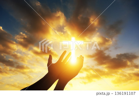 Human hands open palm up worshiping a glowing Christian cross in sunset sky. Concept for faith, spirituality, and hope. 136191107