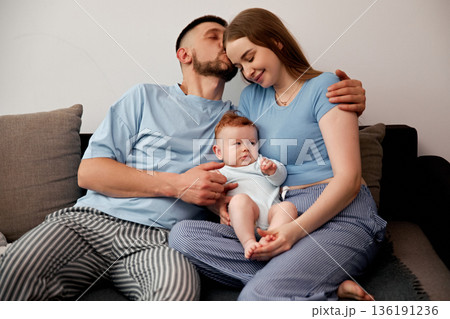 Loving family moment with parents and baby sitting together on a couch, showcasing warmth and affection in a cozy home environment, family bonding concept 136191236
