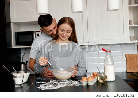 Couple cooking together in a cozy kitchen while preparing batter with joyful expressions and flour scattered on the counter 136191272