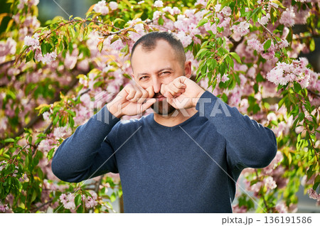 Man allergic suffering from seasonal allergy at spring in blossoming garden at springtime. Bearded young man sneezing and having runny nose in front of blooming tree. Spring allergy concept. 136191586