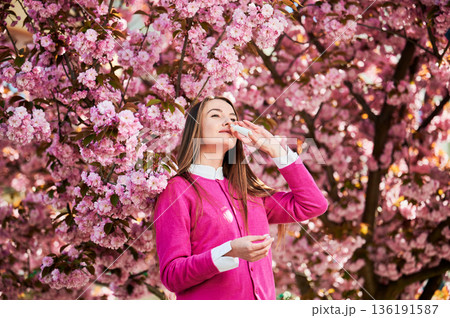Woman allergic using medical nasal spray, suffering from seasonal allergy at spring in blossoming garden. Young woman treating runny nose in front of blooming tree outdoors. Spring allergy concept. Woman allergic using medical nasal spray, suffering from seasonal allergy at spring in blossoming garden. Young woman treating runny nose in front of blooming tree outdoors. Spring allergy concept. 136191587