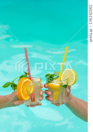 Two hands hold colorful cocktails garnished with fruits and mint over a blue pool. Perfect for depicting leisure, summer relaxation, and tropical vacation vibes. Two hands hold colorful cocktails garnished with fruits and mint over a blue pool. Perfect for depicting leisure, summer relaxation, and tropical vacation vibes. 136192002