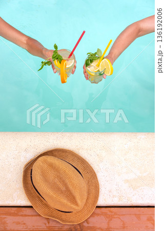 Two hands hold colorful cocktails garnished with fruits and mint over a blue pool. A straw hat rests on the pool edge. Perfect for depicting leisure, summer relaxation, and tropical vacation vibes. Two hands hold colorful cocktails garnished with fruits and mint over a blue pool. A straw hat rests on the pool edge. Perfect for depicting leisure, summer relaxation, and tropical vacation vibes. 136192006