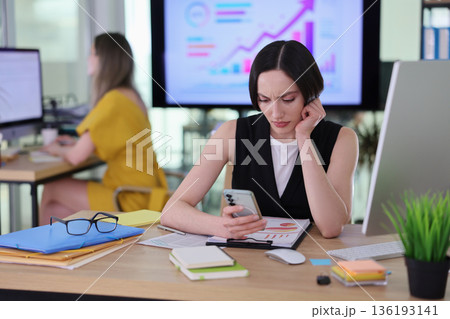 Busy office scene with focused woman checking mobile phone among papers and reports 136193141