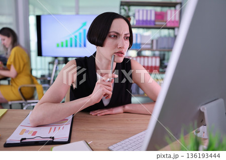 Focused business woman analyzing data in a modern office while another colleague works on a project in the background Focused business woman analyzing data in a modern office while another colleague works on a project in the background 136193444