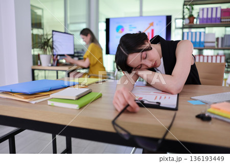 Busy office scene shows hardworking woman feeling exhausted while her colleague focuses on computer tasks Busy office scene shows hardworking woman feeling exhausted while her colleague focuses on computer tasks 136193449