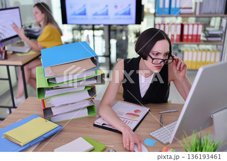 A focused professional works at a cluttered desk with data charts while a colleague collaborates in the busy background A focused professional works at a cluttered desk with data charts while a colleague collaborates in the busy background 136193461
