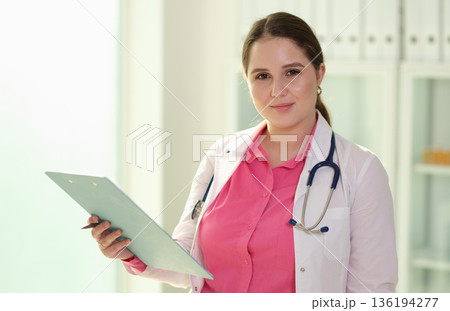 Confident female doctor in a modern clinic holds patient chart ready for consultation in bright, welcoming environment 136194277