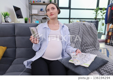 Expecting mother contemplates her future while surrounded by cash in a cozy living room setting Expecting mother contemplates her future while surrounded by cash in a cozy living room setting 136194294