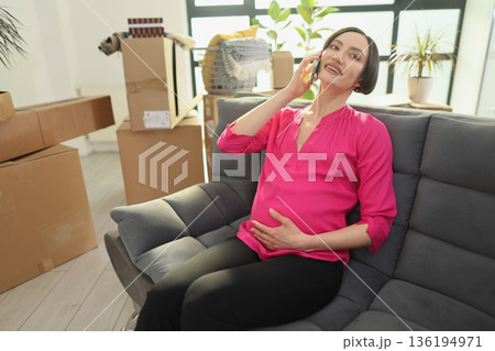 Pregnant woman talks joyfully on the phone while sitting on a cozy couch surrounded by moving boxes Pregnant woman talks joyfully on the phone while sitting on a cozy couch surrounded by moving boxes 136194971