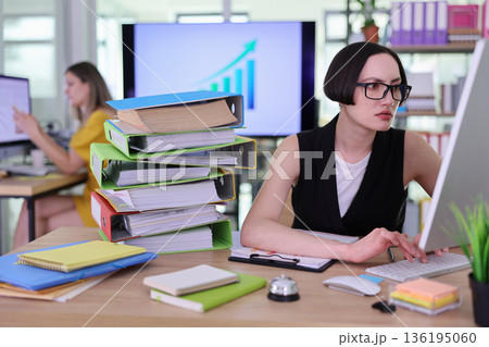 A dedicated worker sits at a desk, typing intently on a keyboard among stacks of vibrant file folders A dedicated worker sits at a desk, typing intently on a keyboard among stacks of vibrant file folders 136195060
