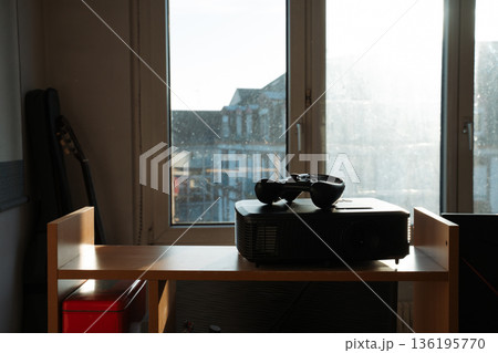Game controller resting on top of a projector on a wooden shelf in sunlight. Casual home interior scene showing gaming setup. Concept of home, technology, gaming, leisure, lifestyle, entertainment. Game controller resting on top of a projector on a wooden shelf in sunlight. Casual home interior scene showing gaming setup. Concept of home, technology, gaming, leisure, lifestyle, entertainment. 136195770