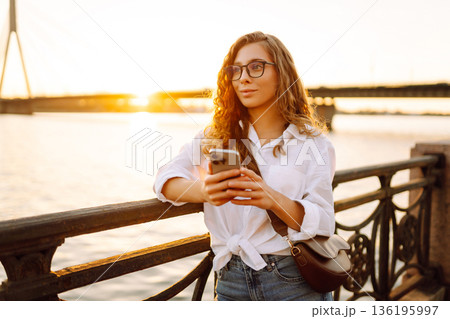 Young woman uses phone walk on embankment. Concept of vacation, technology, weekend. Young woman uses phone walk on embankment. Concept of vacation, technology, weekend. 136195997