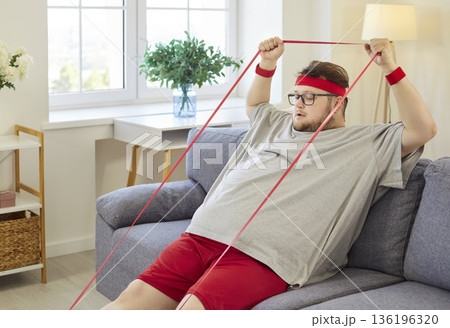 Overweight man doing funny home workout with resistance band on living room sofa 136196320