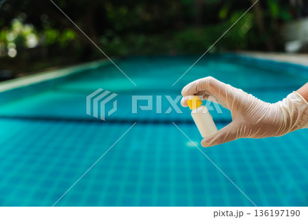 Hand in protective glove holding small test reagent bottle above turquoise swimming pool, water quality control and pool care concept with tropical background Hand in protective glove holding small test reagent bottle above turquoise swimming pool, water quality control and pool care concept with tropical background 136197190