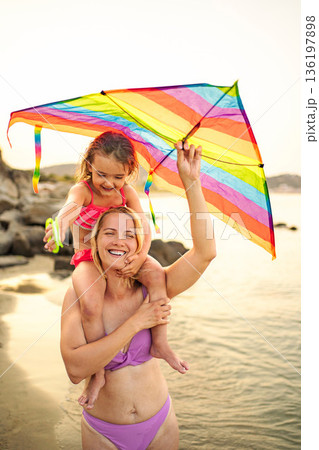 Happy mother carrying daughter on shoulders playing kite at beach 136197898