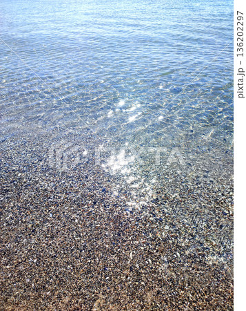 Clean transparent water on shallow sandy seashore with shells with shells on sunny summer day. Sun glare on surface of water. Small waves on sea coast. Nature backdrop. Natural background Clean transparent water on shallow sandy seashore with shells with shells on sunny summer day. Sun glare on surface of water. Small waves on sea coast. Nature backdrop. Natural background 136202297