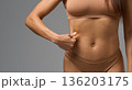 Close-up of woman in beige underwear pinching skin on side of stomach in studio light. 136203175
