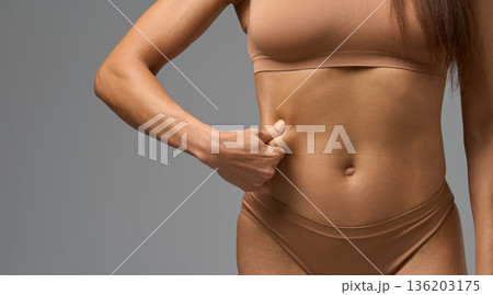 Close-up of woman in beige underwear pinching skin on side of stomach in studio light. Close-up of woman in beige underwear pinching skin on side of stomach in studio light. 136203175