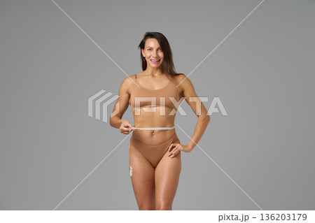 Full-length portrait of fit woman measuring waist with tape showing confidence and body positivity 136203179