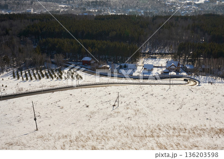 Aerial view of a winding road in a snowy winter landscape with forest, houses and village in the distance for travel and nature concept. Aerial view of a winding road in a snowy winter landscape with forest, houses and village in the distance for travel and nature concept. 136203598