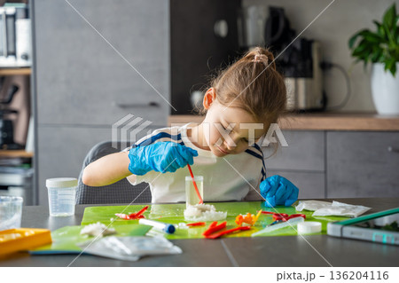 Young girl stirring chemical solution with measuring spoon in cup during crystal growing experiment at home. Chemistry practice and early STEM education through practical laboratory activity. Young girl stirring chemical solution with measuring spoon in cup during crystal growing experiment at home. Chemistry practice and early STEM education through practical laboratory activity. 136204116