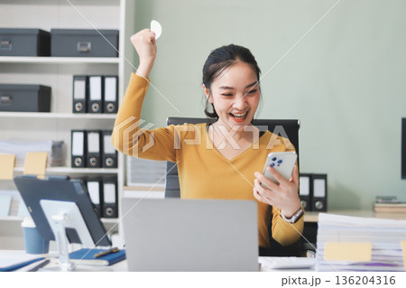 Asian woman works on laptop at office desk. She smiles happily while typing on keyboard near window with blinds. Professional woman works in business environment. 136204316