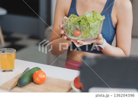 Fit young Asian women in activewear sitting on yoga mat and eating fresh vegetable salad after workout, representing clean eating, fitness lifestyle, wellness routine and health balance. 136204396
