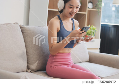 Fit young Asian women in activewear sitting on yoga mat and eating fresh vegetable salad after workout, representing clean eating, fitness lifestyle, wellness routine and health balance. 136206912