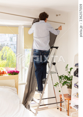 A man is using a ladder to install curtains on a rod in a well-lit room. He holds gray curtains in one hand while adjusting the rod above a window. Plants and flowers are seen outside A man is using a ladder to install curtains on a rod in a well-lit room. He holds gray curtains in one hand while adjusting the rod above a window. Plants and flowers are seen outside 136207009